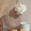 Fleur de bain écologique en fibre de bambou coloris crème, suspendue devant un mur en carreaux de terre cuite avec un savon artisanal.