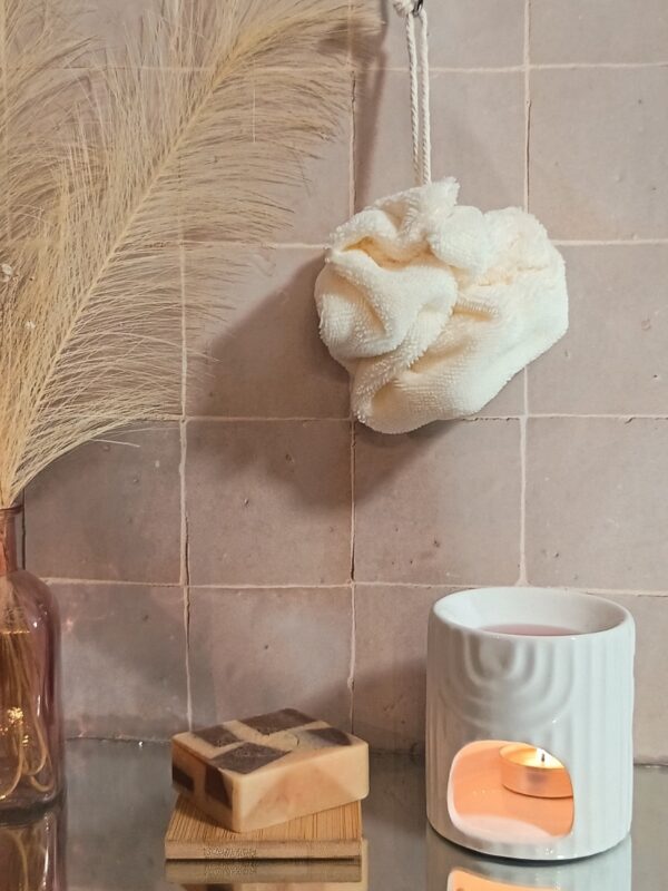 Fleur de bain écologique en fibre de bambou coloris crème, suspendue devant un mur en carreaux de terre cuite avec un savon artisanal.