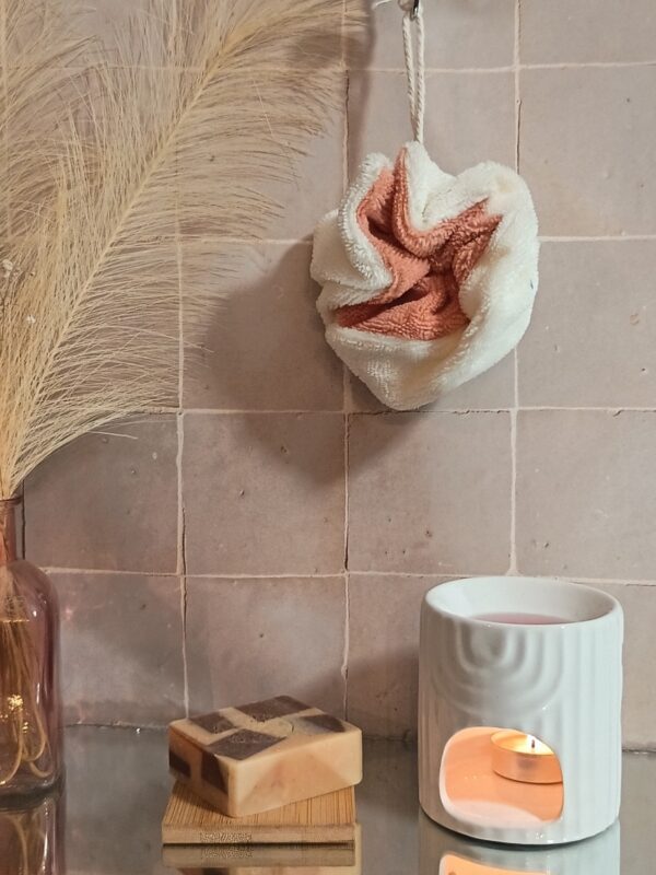 Fleur de douche bicolore pêche et crème en bambou, suspendue devant un mur en carreaux de terre cuite avec un savon artisanal.