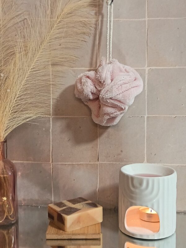 Fleur de bain éco-responsable coloris rose poudré, suspendue par sa cordelette blanche dans un décor de bien-être.