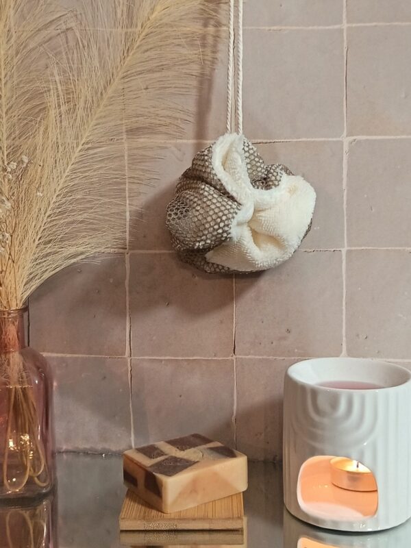 Fleur de douche lavable bicolore crème et taupe suspendue devant un mur en carrelage rose poudré, à côté d'un vase de pampas et d'un brûle-parfum.