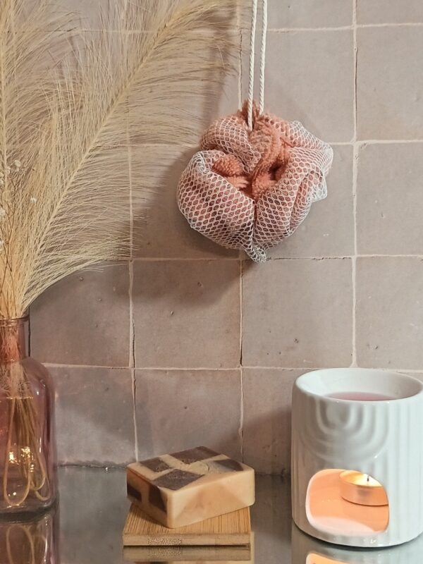 Fleur de douche réutilisable coloris pêche et filet blanc, suspendue au-dessus d'un savon artisanal posé sur un support en bambou.