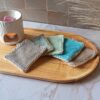 Assortiment de lingettes exfoliantes lavables aux couleurs naturelles (gris, vert d'eau, turquoise, taupe) disposées sur un plateau en bois à côté d'un brûle-parfum blanc. différente couleur