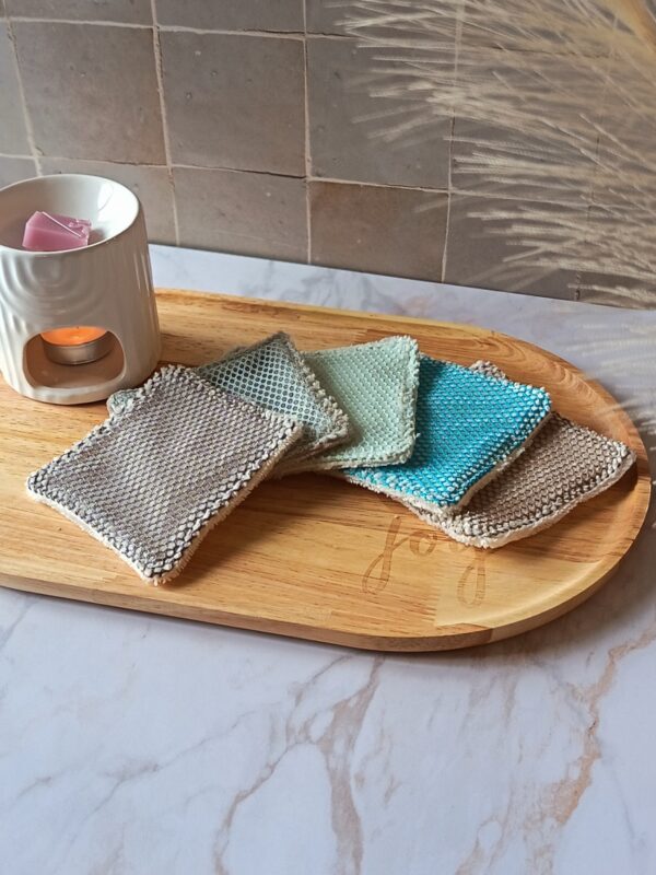 Assortiment de lingettes exfoliantes lavables aux couleurs naturelles (gris, vert d'eau, turquoise, taupe) disposées sur un plateau en bois à côté d'un brûle-parfum blanc. différente couleur