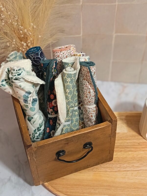 Boîte en bois vintage contenant plusieurs serviettes pour le visage en bambou roulées, laissant apparaître des motifs variés : feuilles d'eucalyptus, hexagones géométriques verts et fleurs colorées.