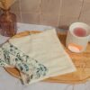 Vue de dessus d'une serviette visage en bambou dépliée de couleur crème, posée sur un plateau en bois à côté d'une serviette roulée avec un motif de feuilles d'eucalyptus bleues et vertes.