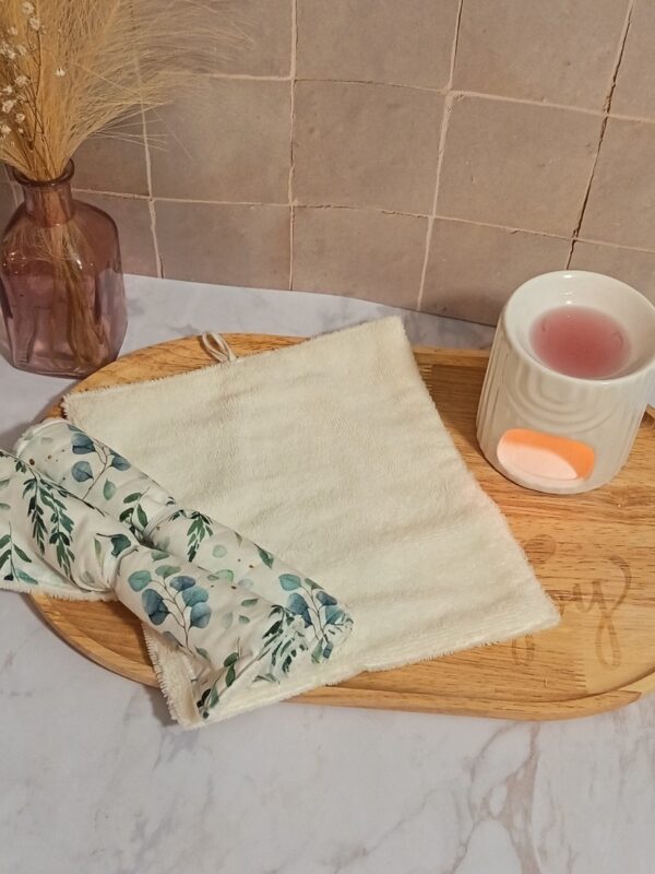 Vue de dessus d'une serviette visage en bambou dépliée de couleur crème, posée sur un plateau en bois à côté d'une serviette roulée avec un motif de feuilles d'eucalyptus bleues et vertes.