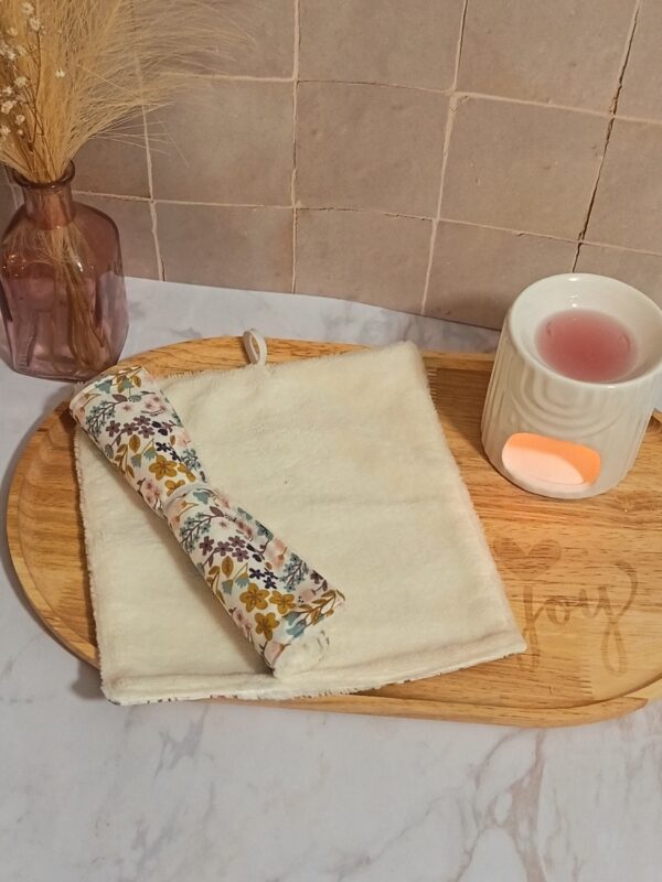 Serviette visage en fibre de bambou crème avec son revers en tissu fleuri "Milly" aux tons ocre, bleu et violet. La serviette est présentée roulée sur un plateau en bois dans une ambiance spa.