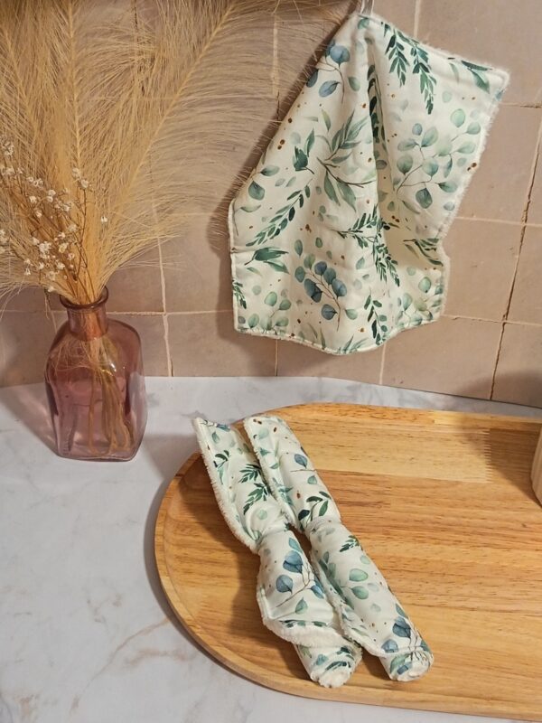 Serviette visage en bambou suspendue par sa petite boucle blanche, révélant un tissu imprimé de feuilles d'eucalyptus. Deux autres serviettes assorties sont enroulées sur un plateau en bois à côté.