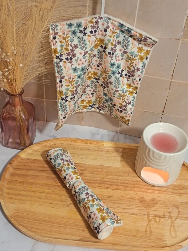 Petite serviette de toilette multi-usage suspendue au mur, affichant un motif fleuri coloré "Milly". Une serviette enroulée est posée sur un plateau décoratif avec un brûle-parfum.