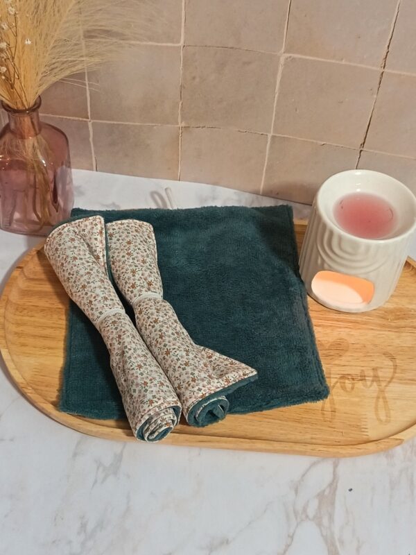 Serviette visage en éponge de bambou coloris indigo (vert canard foncé) posée à plat, surmontée de deux serviettes enroulées au motif "Mini Pio" avec de petites fleurs ocre et roses.