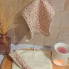Serviette visage en éponge bambou crème suspendue, mettant en avant un tissu à micro-fleurs ocre et violettes. Une version enroulée est posée sur un plateau en bois à côté d'un diffuseur.