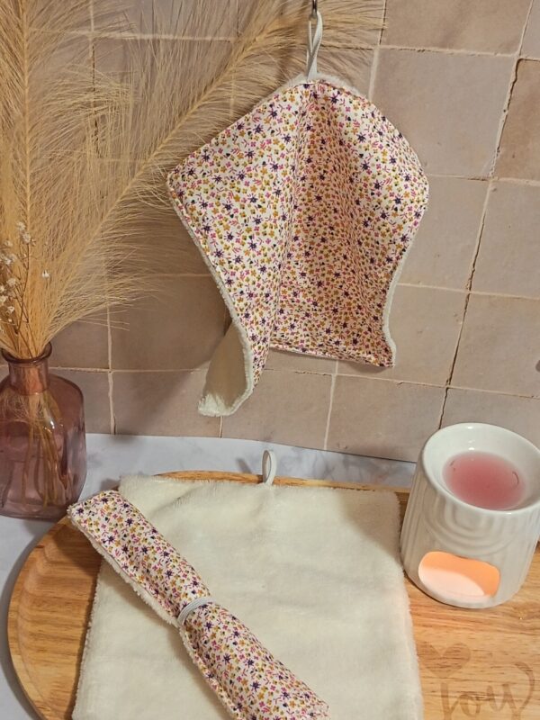 Serviette visage en éponge bambou crème suspendue, mettant en avant un tissu à micro-fleurs ocre et violettes. Une version enroulée est posée sur un plateau en bois à côté d'un diffuseur.