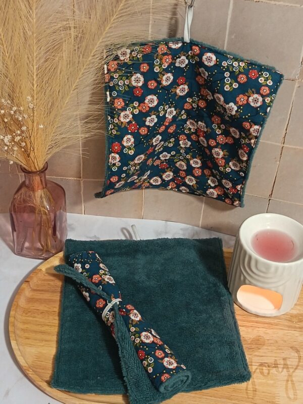 Carré de soin suspendu présentant le motif "Okaido" (fleurs japonaises) sur une éponge bambou bleu pétrole. Une serviette est aussi présentée enroulée sur le plateau "Joy".