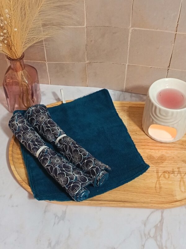 Duo de serviettes visage enroulées avec motif floral bleu marine et blanc, posées sur une face éponge en bambou bleu pétrole dépliée.