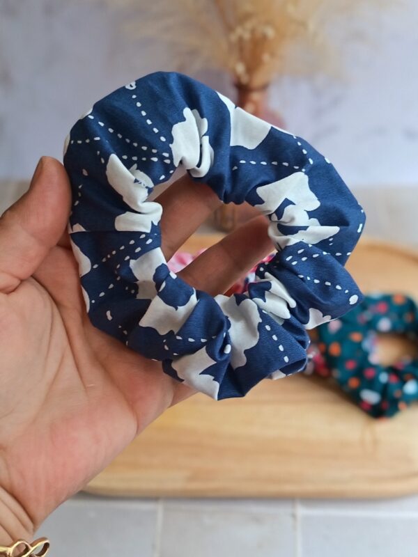 Chouchou à cheveux en coton bleu avec fleur blanche, finitions soignées, tenu entre les doigts.