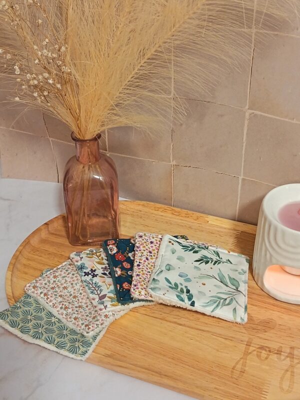 Éventail de lingettes démaquillantes lavables avec différents motifs fleuris et géométriques, disposé sur un plateau en bois dans une ambiance cocooning avec un vase de fleurs séchées.