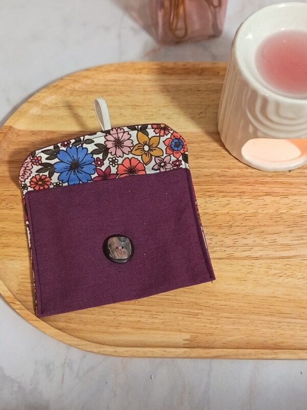 Vue de face de l'étui à savon violet modèle Brigitte, ouvert. Le rabat révèle une doublure intérieure en tissu fleuri coloré (bleu, jaune, orange). Un élastique blanc sert de fermeture sur un bouton central noir.