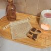 Un filet à savon vide en coton naturel posé sur un plateau en bois clair, à côté d'un savon artisanal bicolore. En arrière-plan, une décoration zen avec un petit vase et un diffuseur de parfum blanc.