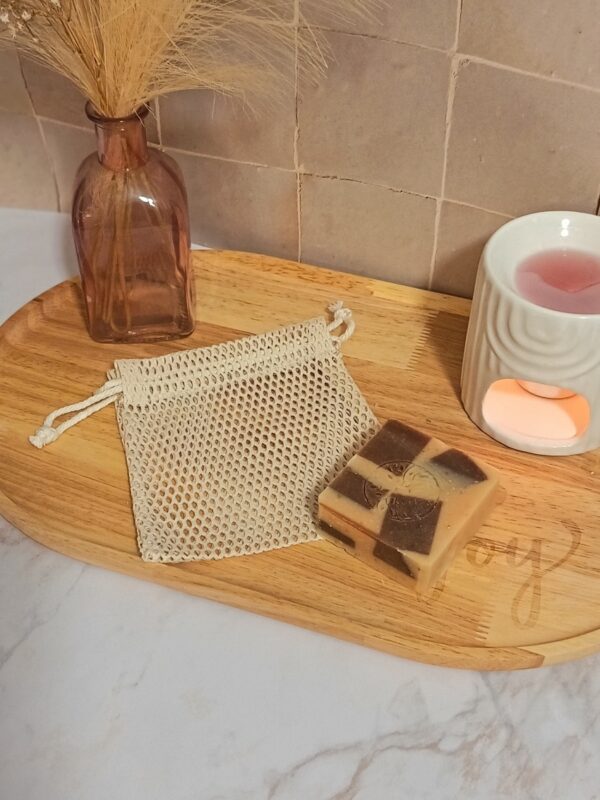 Un filet à savon vide en coton naturel posé sur un plateau en bois clair, à côté d'un savon artisanal bicolore. En arrière-plan, une décoration zen avec un petit vase et un diffuseur de parfum blanc.