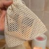 Gros plan d'une main tenant un filet à savon en coton à mailles serrées. Un savon est inséré à l'intérieur, laissant deviner sa texture à travers les mailles beiges.