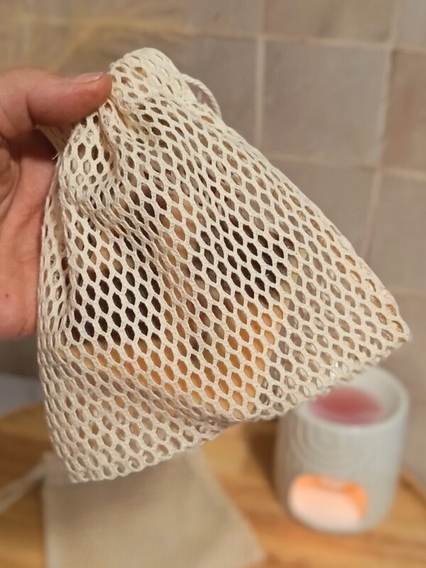 Gros plan d'une main tenant un filet à savon en coton à mailles serrées. Un savon est inséré à l'intérieur, laissant deviner sa texture à travers les mailles beiges.