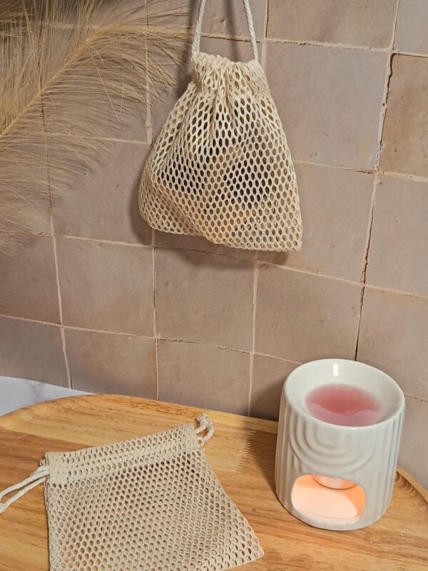 Un filet à savon rempli et suspendu par son cordon contre un mur en carrelage beige. En dessous, un autre filet vide repose sur un plateau en bois à côté d'un brûle-parfum allumé.
