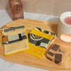 Deux gants sauve-savon artisanaux avec un motif géométrique abstrait gris et jaune moutarde, l'un avec une face en filet de coton et l'autre en tissu éponge, posés sur un plateau en bois à côté d'un savon solide.