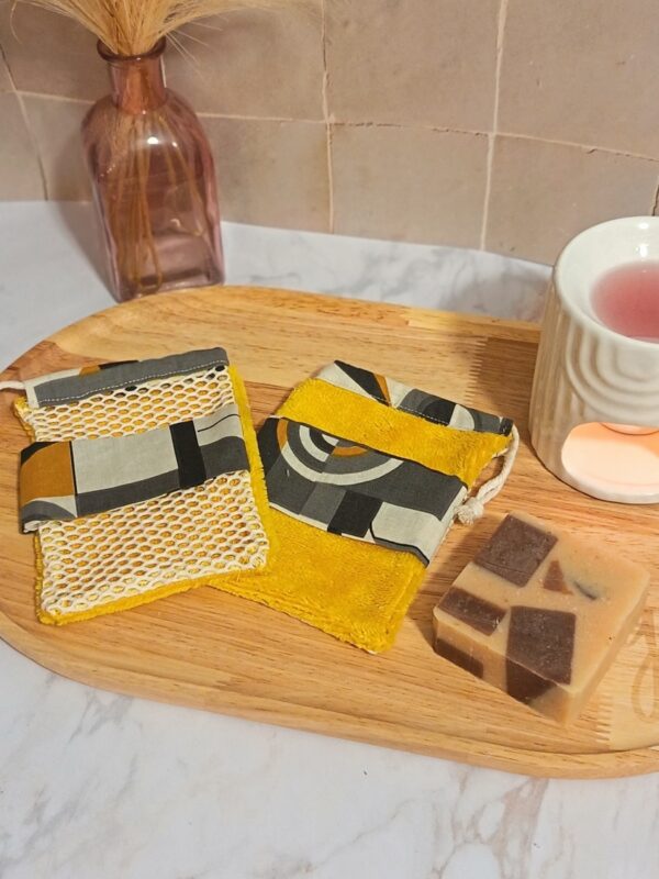 Deux gants sauve-savon artisanaux avec un motif géométrique abstrait gris et jaune moutarde, l'un avec une face en filet de coton et l'autre en tissu éponge, posés sur un plateau en bois à côté d'un savon solide.