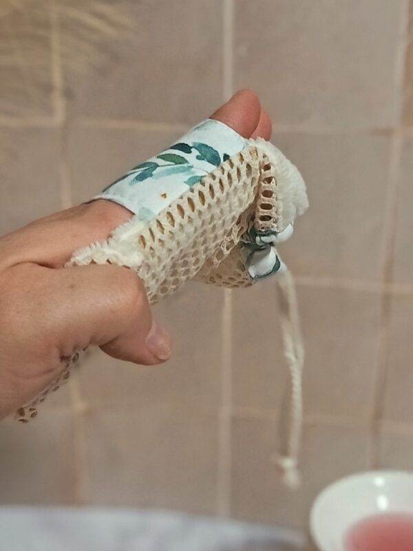 Zoom sur un gant sauve-savon porté à la main, montrant la texture alvéolée du filet en coton bio et une bande de tissu aux motifs de feuilles d'eucalyptus.