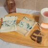 Ensemble de deux gants sauve-savon artisanaux avec motifs eucalyptus et touches dorées, présentés sur un plateau en bois avec un savon solide pour un rituel bain zéro déchet.