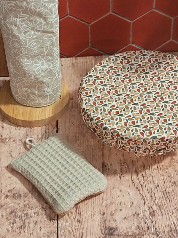 Gros plan sur les accessoires de cuisine éco-responsables : éponge lavable en nid d'abeille beige, charlotte alimentaire à motifs et essuie-tout réutilisable sur support en bois.