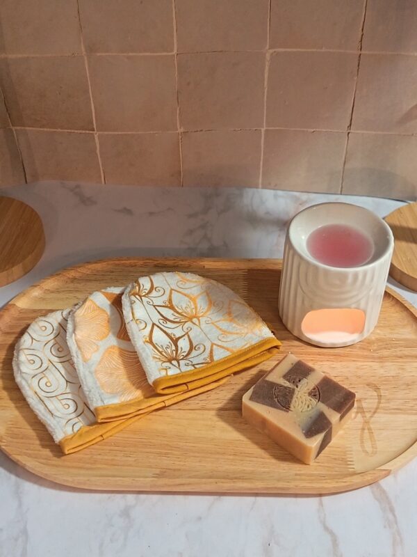 Lot de trois mini-gants de toilette blancs avec des motifs graphiques et floraux dorés, posés sur un plateau en bois à côté d'un savon artisanal et d'un brûle-parfum blanc.