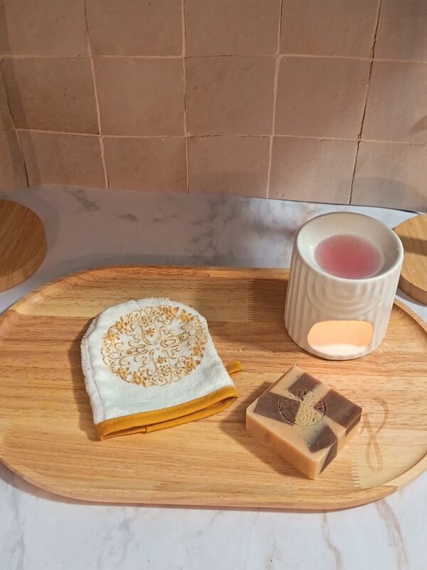 Mini-gant de toilette blanc posé sur un plateau en bois, orné d'un médaillon floral complexe de couleur or et bordé d'un biais jaune.