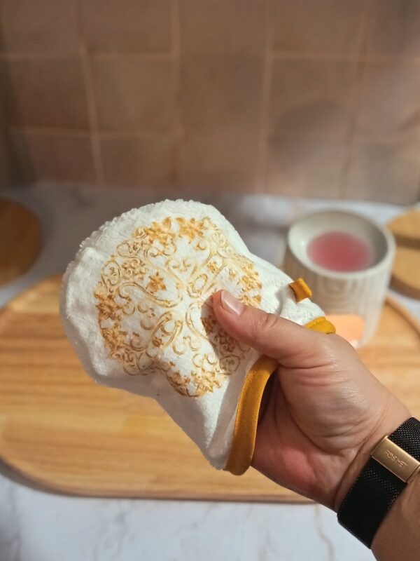 Main présentant un mini-gant en bambou blanc avec un motif de rosace dorée élégante, idéal pour le soin du visage.
