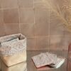 Panier de rangement en tissu et simili-cuir beige contenant des lingettes lavables assorties au motif floral Mini Pio, posé sur une surface réfléchissante devant un mur de carreaux artisanaux.