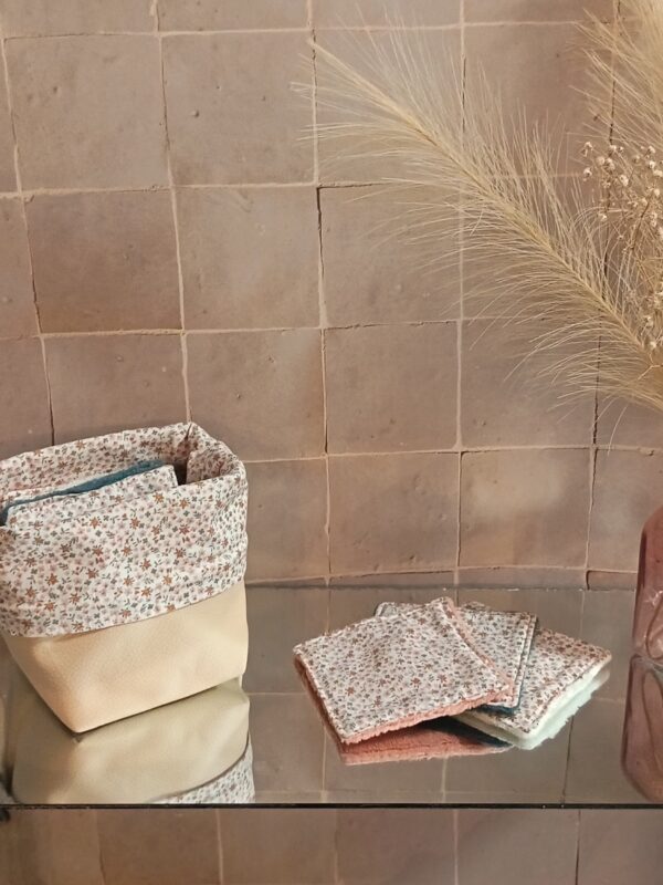 Panier de rangement en tissu et simili-cuir beige contenant des lingettes lavables assorties au motif floral Mini Pio, posé sur une surface réfléchissante devant un mur de carreaux artisanaux.