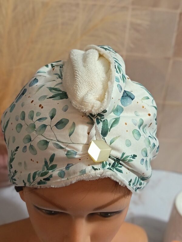 Gros plan sur l'attache d'une serviette à cheveux : élastique blanc fixé sur un bouton hexagonal doré, tissu motif eucalyptus et doublure éponge blanche.