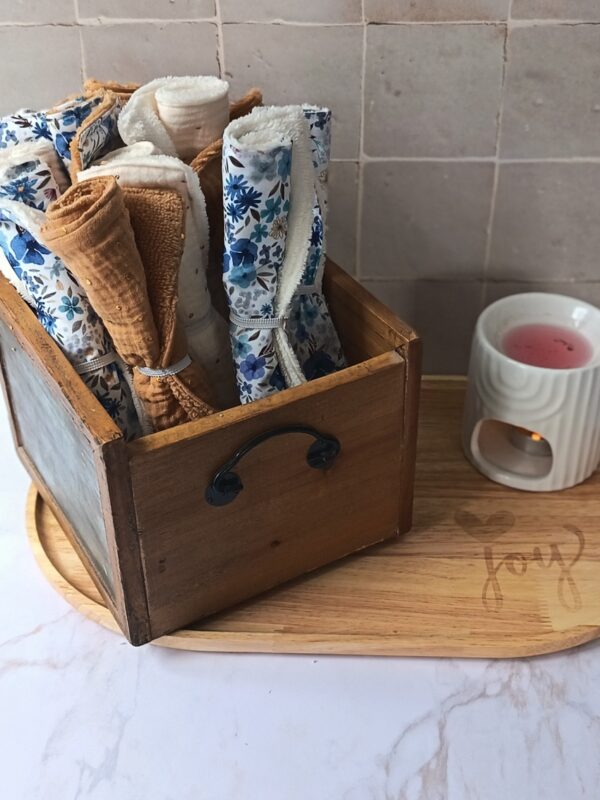 Panier en bois contenant un assortiment de serviettes visage écoresponsables roulées, aux coloris ocre, crème et motifs floraux, présentées dans une ambiance bien-être.