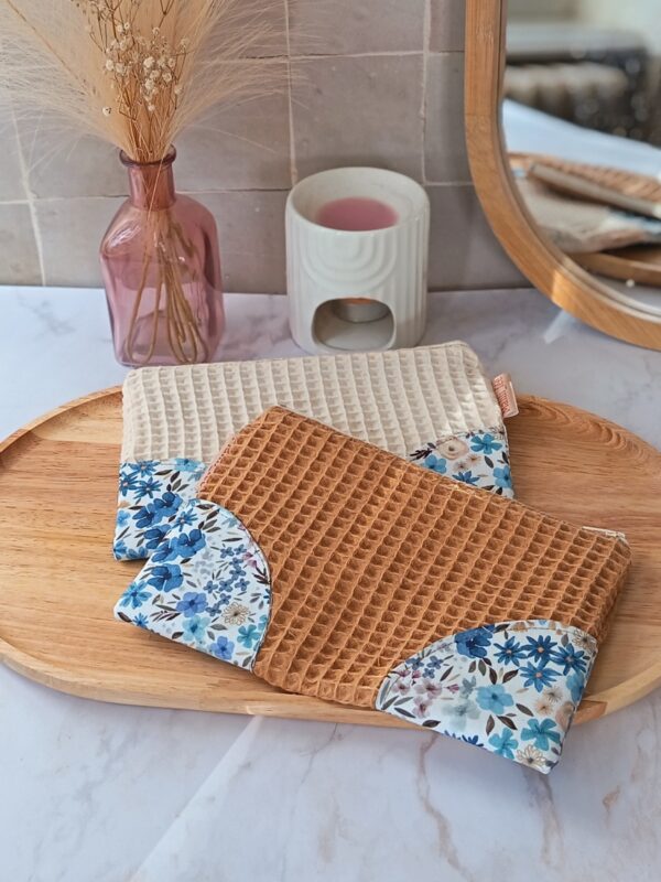 Deux pochettes de rangement artisanales en tissu nid d'abeille ocre et écru avec coins fleuris, posées sur un plateau en bois.