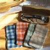 Vue de dessus de quatre essuie-lunettes tissu double gaze artisanaux, aux motifs vichy terracotta, vert, lin et bleu, posés sur un plateau en bois avec un étui à lunettes ouvert.
