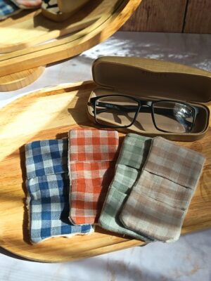 Vue de dessus de quatre essuie-lunettes tissu double gaze artisanaux, aux motifs vichy terracotta, vert, lin et bleu, posés sur un plateau en bois avec un étui à lunettes ouvert.
