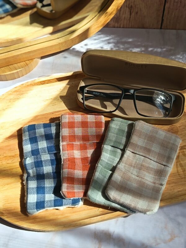 Vue de dessus de quatre essuie-lunettes tissu double gaze artisanaux, aux motifs vichy terracotta, vert, lin et bleu, posés sur un plateau en bois avec un étui à lunettes ouvert.