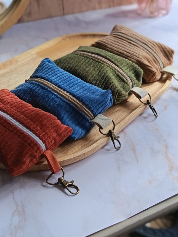 Alignement de quatre étuis nomades en velours côtelé (terracotta, bleu, vert olive, beige) avec mousquetons en bronze, posés sur un plateau en bois.