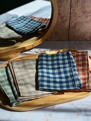 Pile de quatre mouchoirs tissu double gaze artisanaux Malili_fée aux motifs vichy (vert pastel, beige, bleu, terracotta) posée sur un plateau en bois, avec un miroir en arrière-plan.