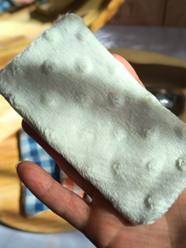 Gros plan montrant la texture douce et texturée de la face en tissu minky blanc d'un essuie-lunettes artisanal, tenu entre les doigts.