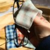 Main tenant un essuie-lunettes en coton vichy beige et vert pour nettoyer une paire de lunettes de vue noires à monture fine.