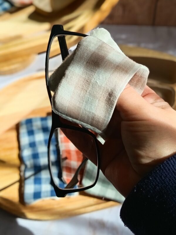 Main tenant un essuie-lunettes en coton vichy beige et vert pour nettoyer une paire de lunettes de vue noires à monture fine.