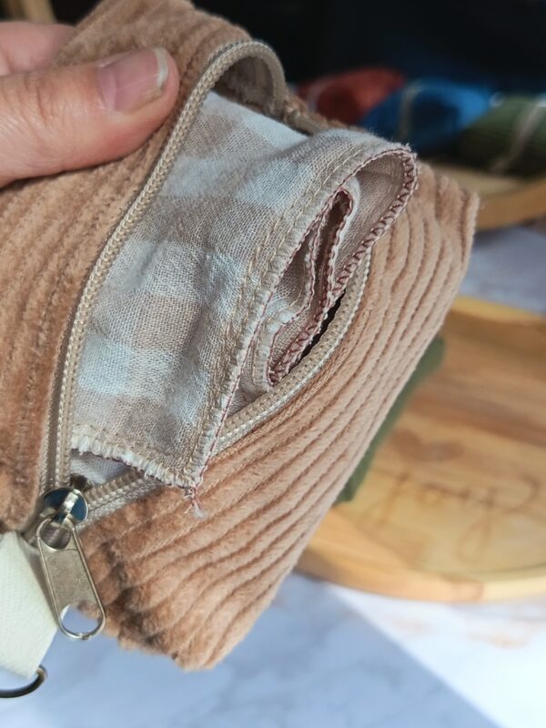 Gros plan montrant un mouchoir tissu double gaze vichy beige inséré dans son étui nomade en velours côtelé beige, avec la fermeture éclair ouverte.