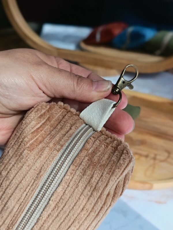 Main tenant le mousqueton en bronze attaché à un étui nomade en velours côtelé Malili_fée, illustrant sa praticité pour l'accrocher partout.